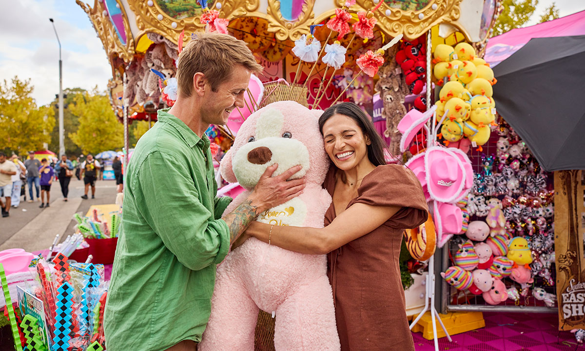 A couple smiling and hugging a large pink teddy bear in front of a colourful game stall at the Sydney Royal Easter Show.