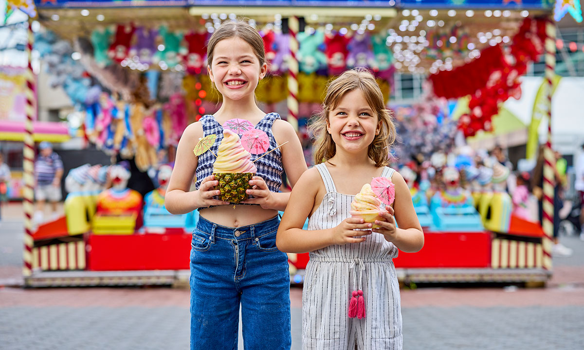 Two young girls smiling while holding colourful ice creams in front of a game stall at the Sydney Royal Easter Show.